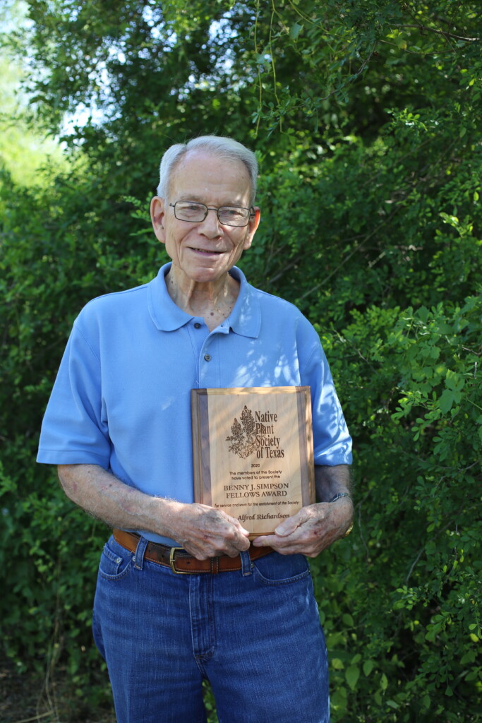 Man in blue button down, holding an award. Standing in front of green foliage.