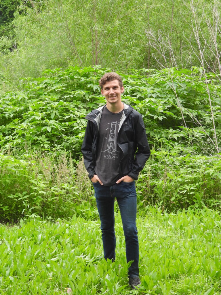 Young person in black jacket and jeans standing in front of green foliage