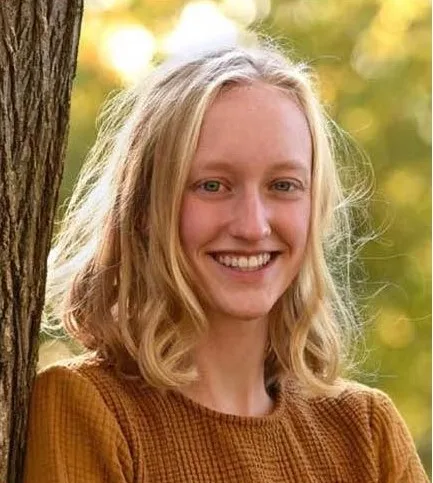 Headshot of a young person with medium length blonde hair; they are leaning up against a tree.